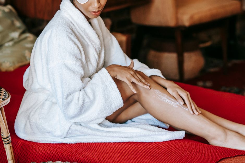 Crop unrecognizable young ethnic female with eye patch applying moisturizing cream on leg while sitting on soft couch